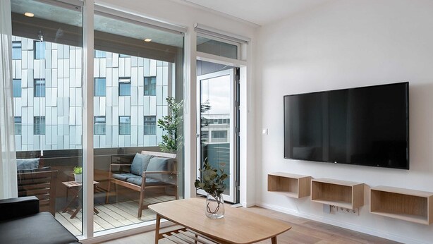 Una sala de estar moderna con un televisor grande, muebles de madera y vistas a un paisaje urbano a través de puertas de cristal amplias.