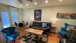 A modern living room with a black leather sofa, two blue armchairs, a wooden coffee table, and a dining area with a glass table and chairs.