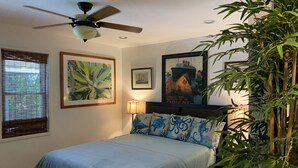 A bedroom with a bed, a ceiling fan, a window with blinds, a framed picture, a lamp, and a large plant.