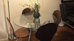 A round glass table with four chairs, a vase with flowers, a floor lamp, and a framed picture on the wall.