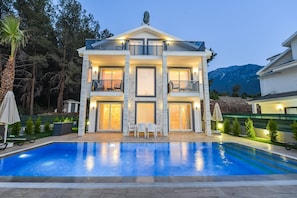 Ein zweistöckiges Haus mit Schwimmbecken davor, umgeben von Bäumen und einem Berg im Hintergrund.