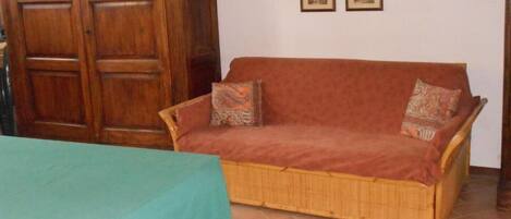 A room with a wooden wardrobe, a wicker sofa with patterned cushions, and a green tablecloth.