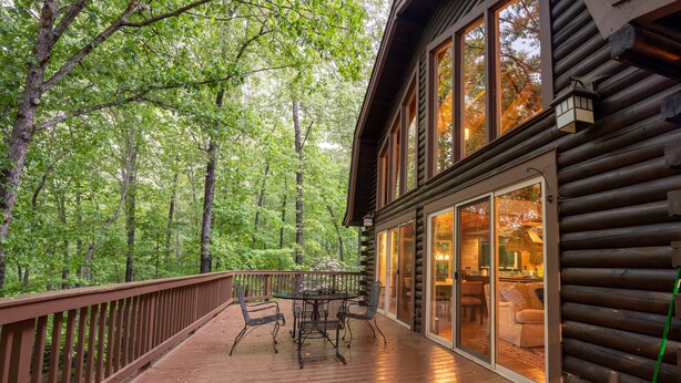 Une terrasse en bois, agrémentée d’une table et de chaises, entourée d’une balustrade en bois, qui mène à un chalet en rondins doté de grandes portes vitrées.