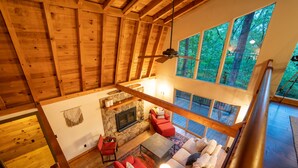 Un vaste espace de vie aménagé dans un style loft, avec une cheminée en pierre, des meubles rouges et blancs, et de grandes fenêtres offrant une vue sur la forêt.