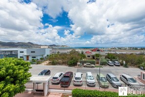 Un parking avec plusieurs voitures, un bâtiment moderne et, au loin, une vue sur la mer et les montagnes.