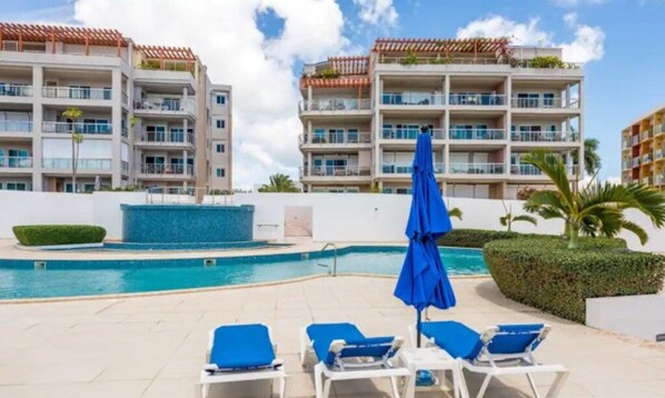 Un espace aménagé au bord de la piscine, avec des chaises longues et un parasol bleu, entouré d’immeubles à plusieurs étages dotés de balcons.