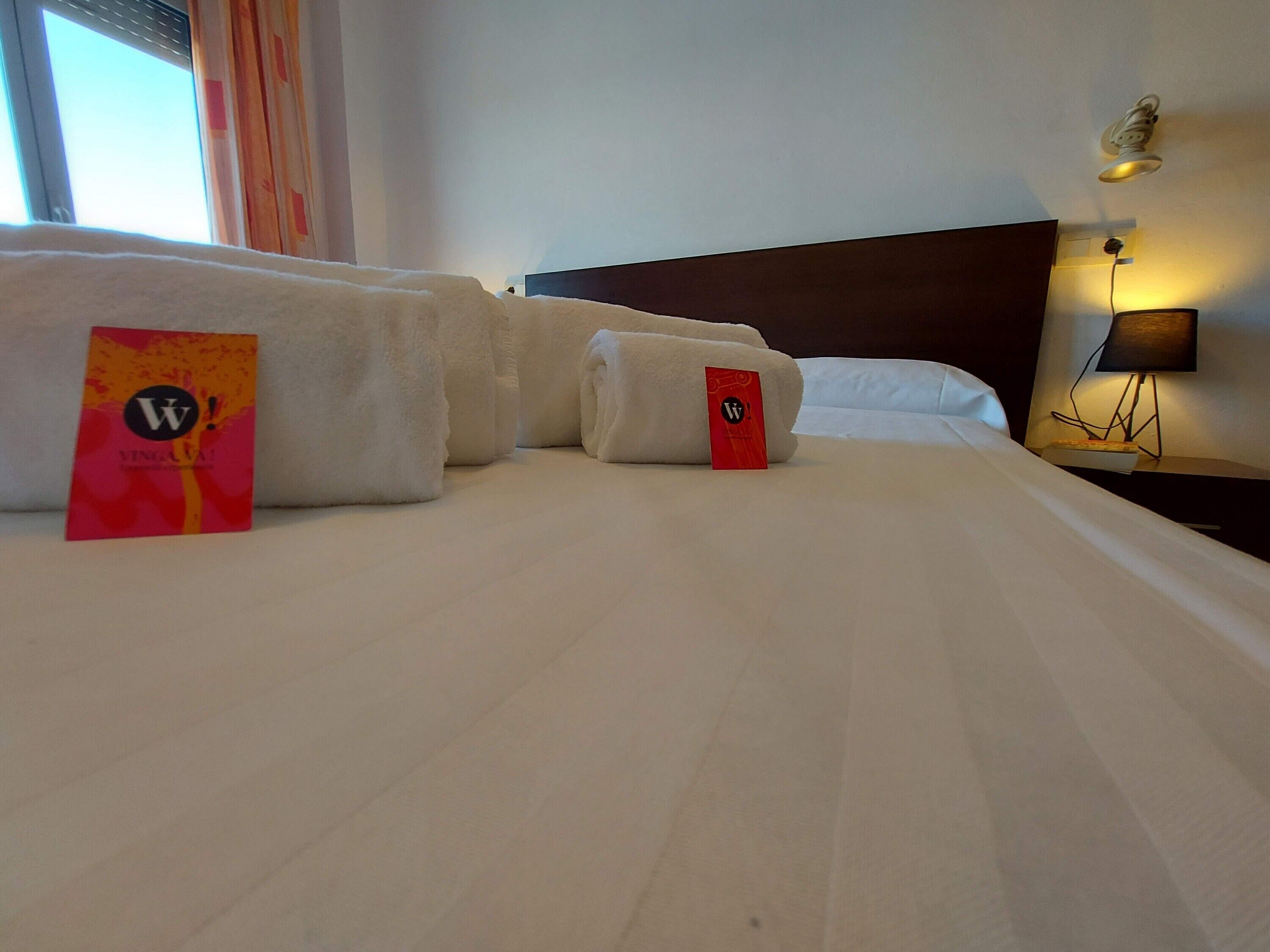 Ein Hotelzimmer mit einem ordentlich bezogenen Bett, einem Nachttisch mit Lampe und einem Fenster mit Vorhängen.