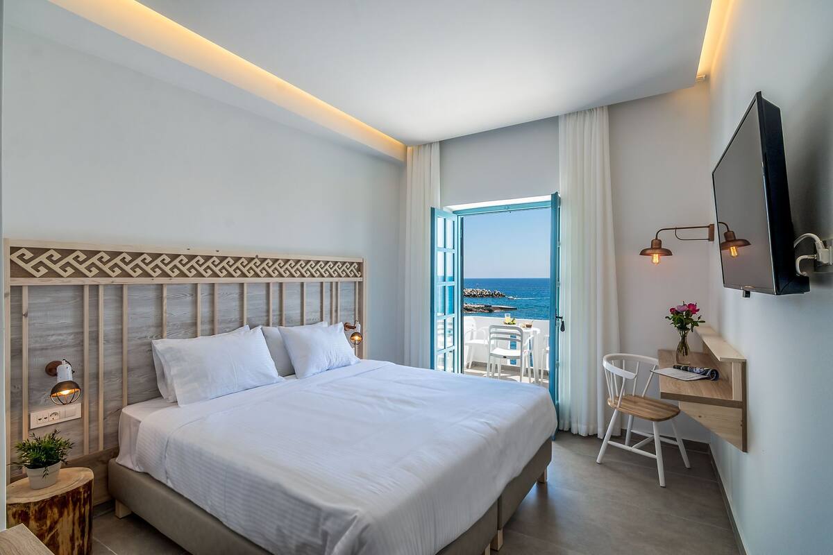A modern hotel room with a large bed, a desk with a chair, and a view of the sea through an open door.