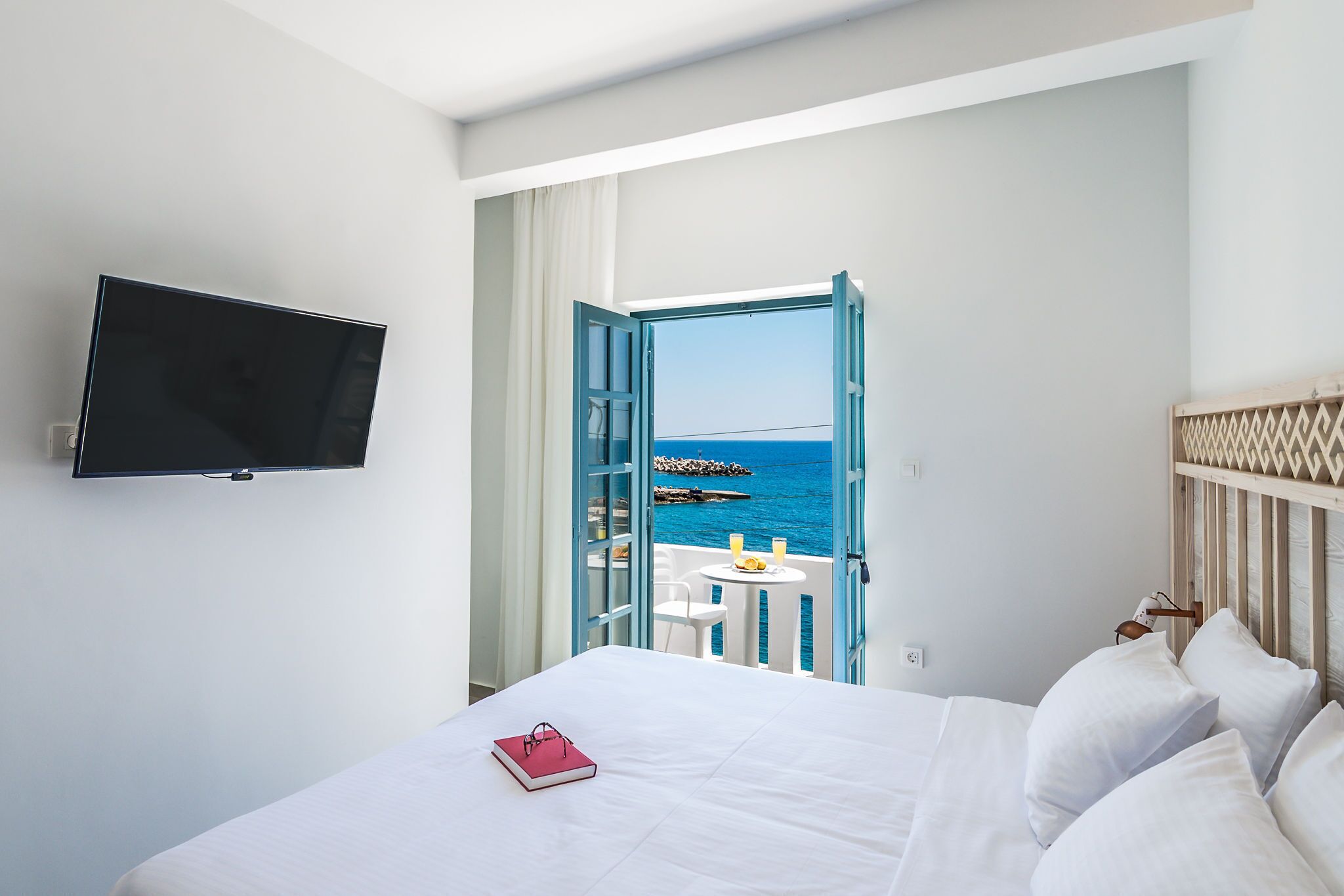 A hotel room with a bed, a TV mounted on the wall, a balcony with a view of the sea, and a small table with a drink.