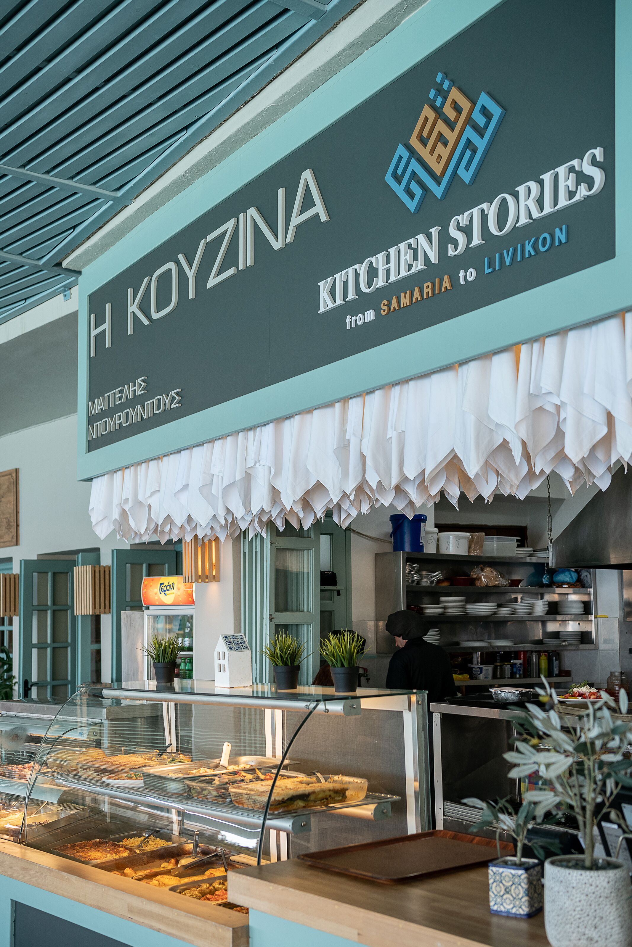 Un restaurante llamado H Koyzina, con un letrero, una vitrina de alimentos y una zona de cocina.