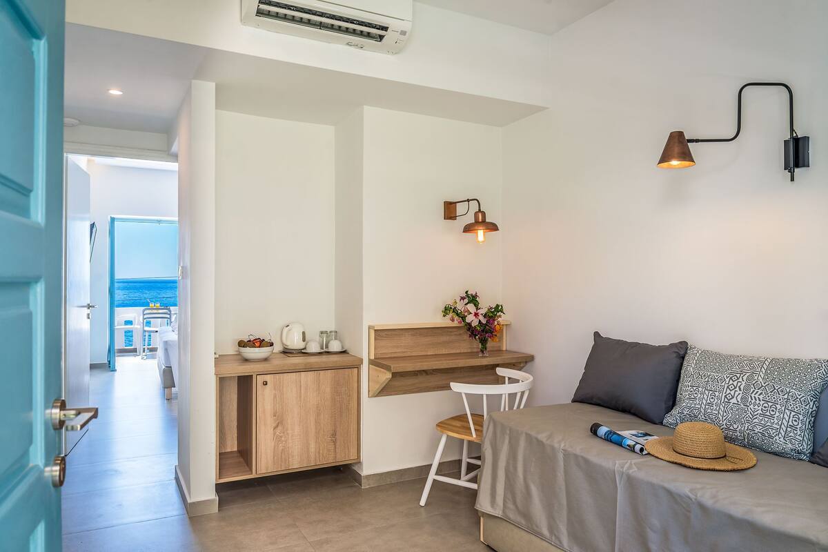 A modern room with a wooden desk, a white chair, a sofa with patterned cushions, a wall-mounted lamp, and a view of the sea through an open door.