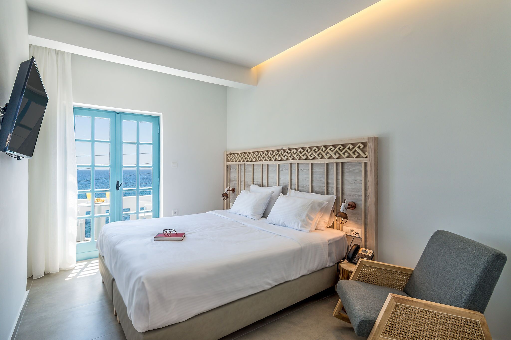 A hotel room with a large bed, a TV mounted on the wall, a wooden headboard, and a view of the sea through a blue door.