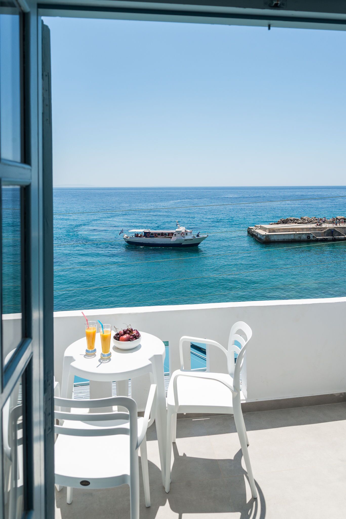 A balcony with a table and chairs, overlooking a calm sea with a boat and a dock.