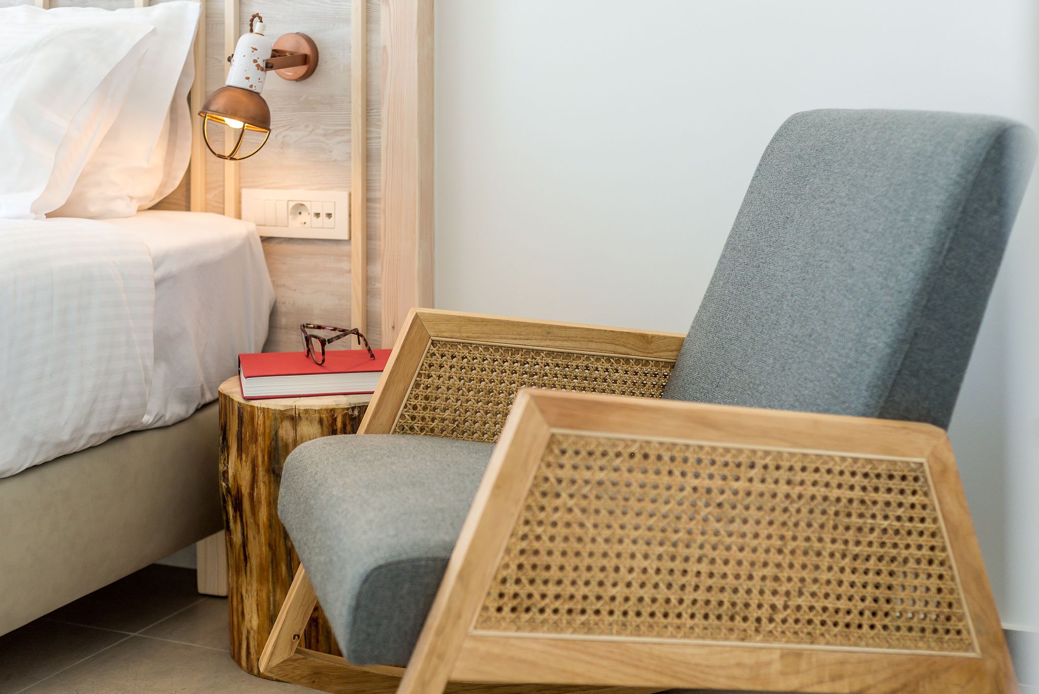 Um quarto com cama, mesa de cabeceira com um livro e óculos, luminária na parede e mesa lateral de madeira com uma cadeira.