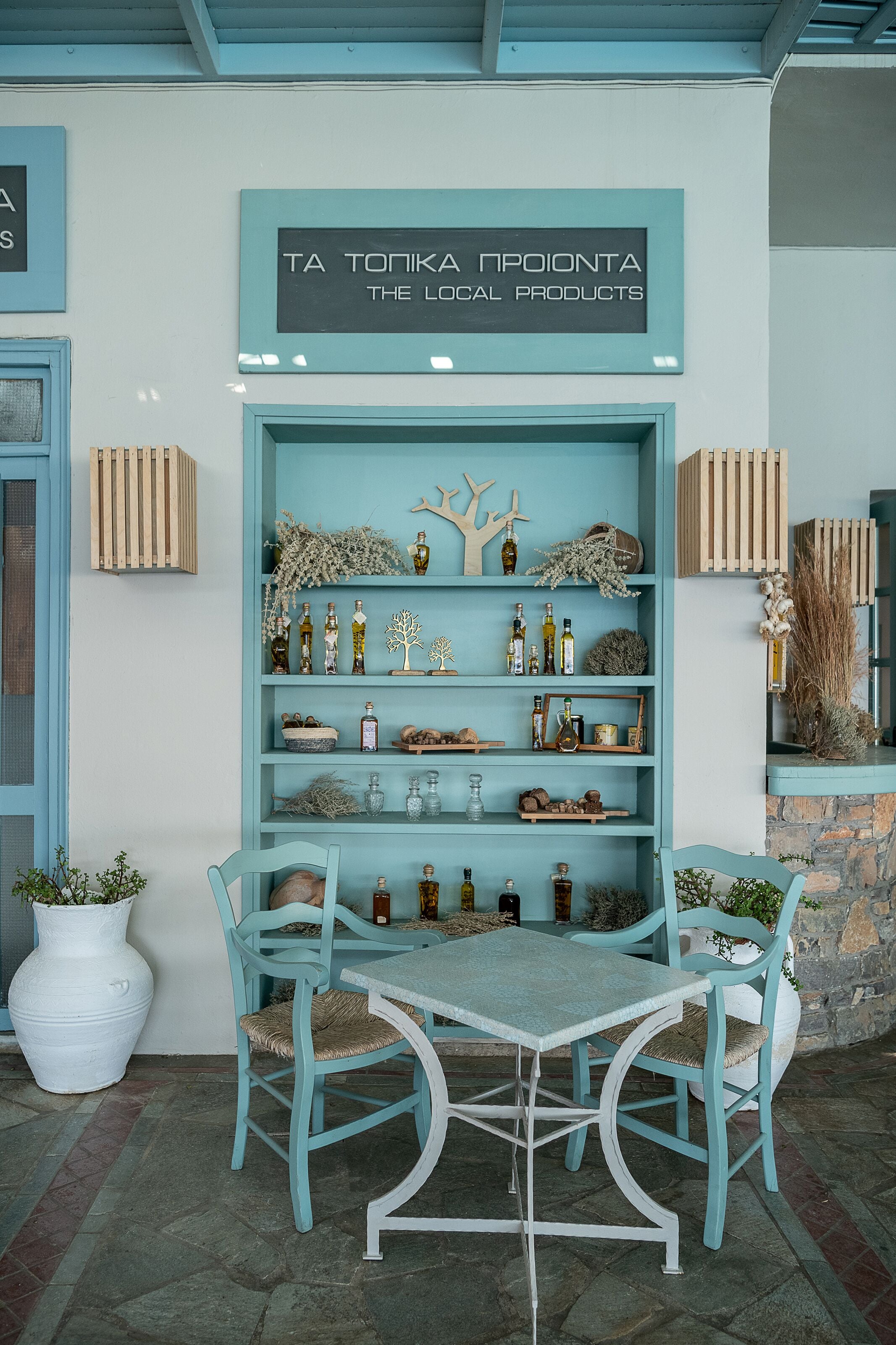 Una estantería azul verdosa con varios objetos, una mesa blanca y dos sillas a juego en una habitación con pared de piedra y un letrero sobre la estantería.