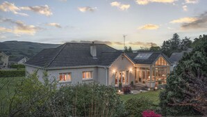 A two-story house with a sloped roof, a visible chimney, and a well-maintained garden with shrubs and flowering plants. - 'The Willows' A Charming home on Dollarbeg Castle Estate (Dollar)