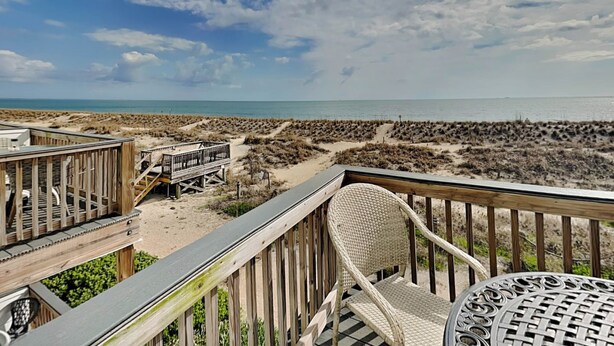 A beach view from a wooden deck with wicker chairs and a table, overlooking the beach and ocean. - Tybee Lights 114C | Pet Friendly, Oceanfront! (Tybee Island)