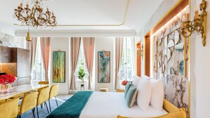 Une chambre d’hôtel avec un lit, un bureau et des chaises. On y trouve des rideaux, un lustre et des œuvres d’art accrochées aux murs.