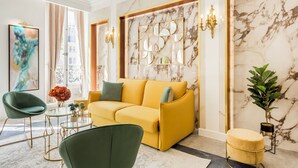 Un salon moderne avec un canapé jaune, des fauteuils verts, une table basse en verre et un mur en marbre agrémenté d’un miroir aux motifs géométriques.