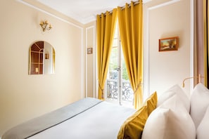 A hotel room with a bed, a window with yellow curtains, a balcony with a wrought iron railing, and a wall-mounted mirror.