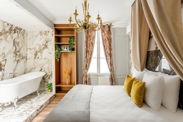 A bedroom with a large bed, a bathtub, a wooden shelf with plants, and a chandelier.
