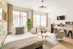 Une chambre d’hôtel moderne avec un grand lit, une petite table, une chaise, un téléviseur et une vue sur la ville grâce à de grandes fenêtres.