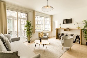 Un salon moderne avec un canapé, une table basse ronde, un fauteuil et un téléviseur fixé au mur. On peut voir des plantes en pot et un balcon à travers les fenêtres.