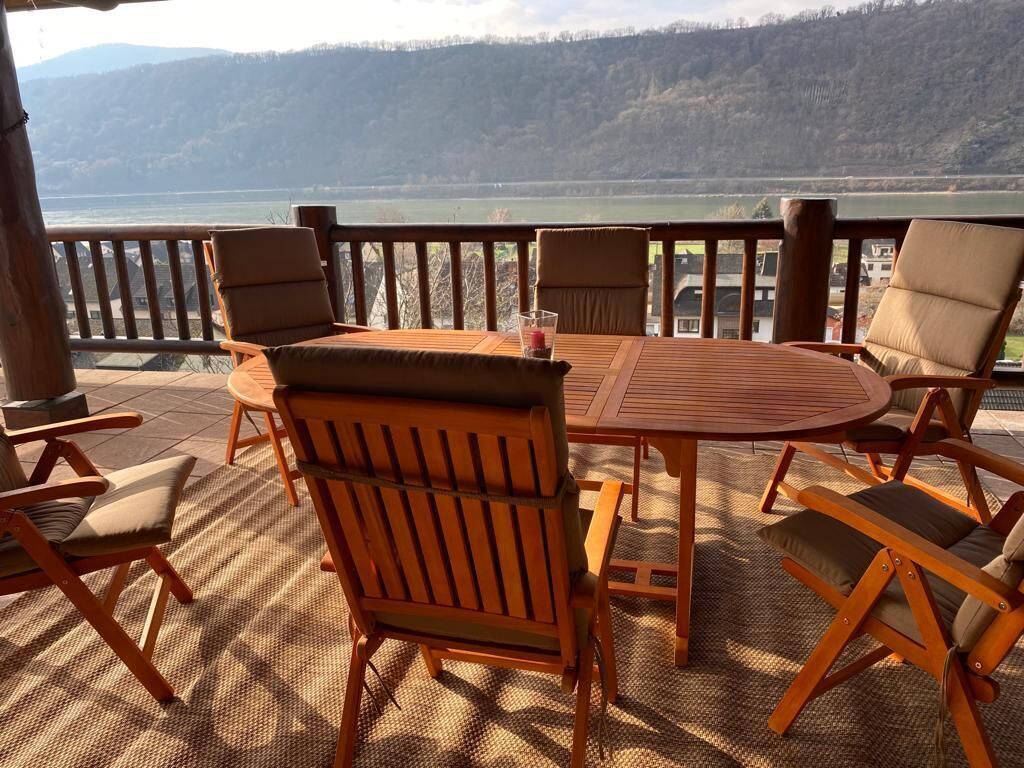 A wooden deck with a round table and chairs, overlooking a lake and hills.