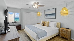 Un dormitorio con cama, mesitas de noche, ventilador de techo, ventana con vista al océano y un cuadro en la pared.