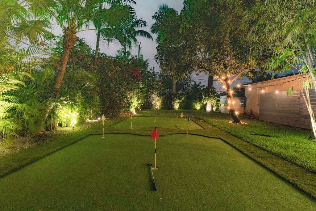 A well-lit mini-golf course with a red flag, surrounded by lush greenery and trees.