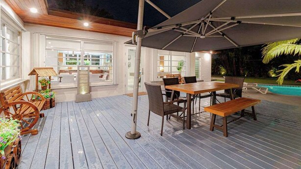 Eine Terrasse im Freien mit Holzdeck, einem Tisch mit Stühlen, einem großen Sonnenschirm und einem Pool im Hintergrund.