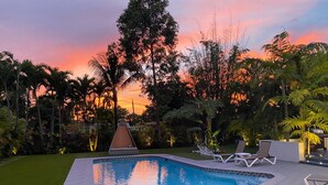 Ein Schwimmbecken mit Rasenfläche, Liegestühlen und Blick auf Bäume und einen Sonnenuntergang.