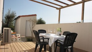 Un patio al aire libre con una mesa lista para comer, rodeado de sillas y una mesita con una planta en maceta y copas de vino.