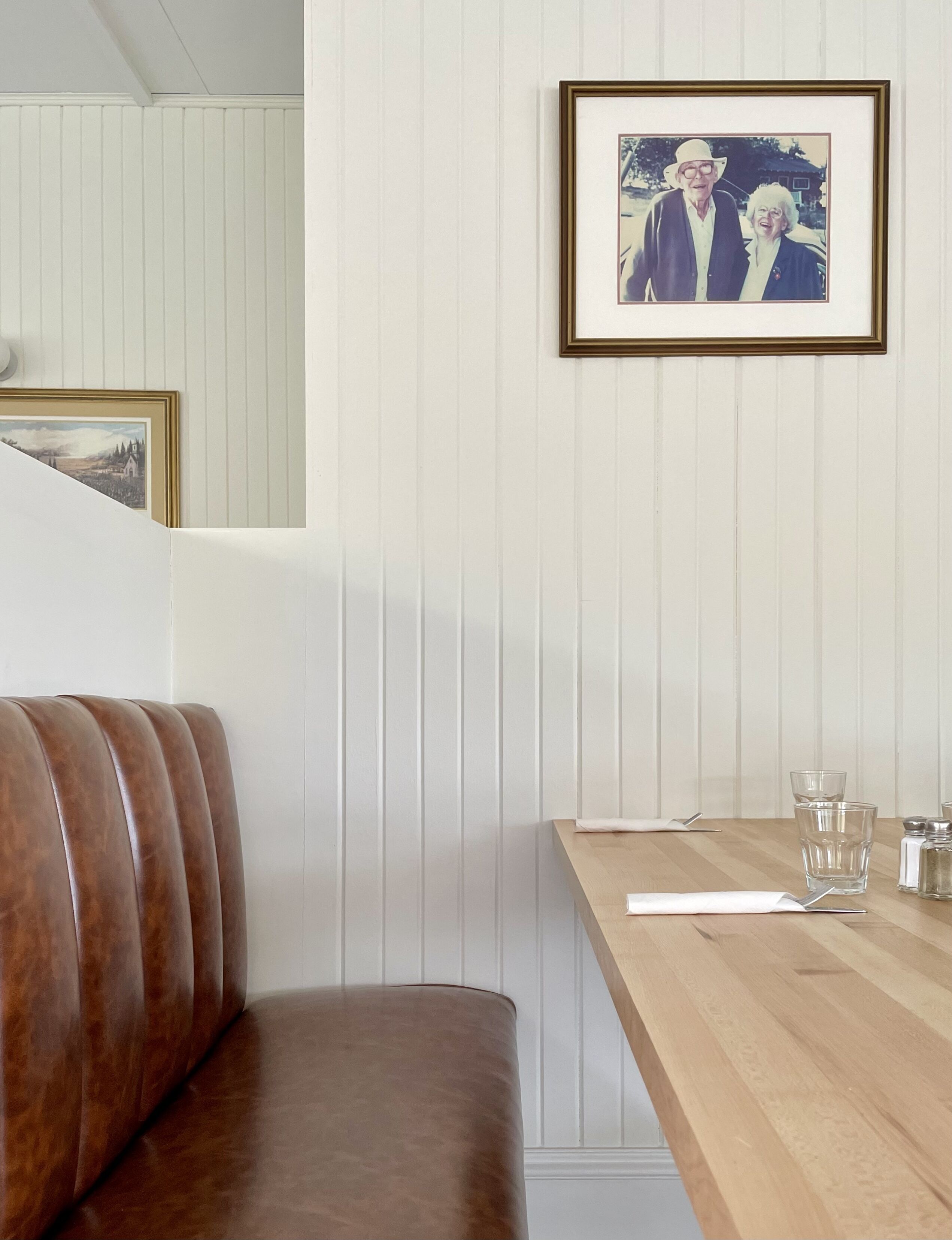 Eine hölzerne Bank mit gepolstertem Sitz, ein gerahmtes Bild an der Wand und ein Holztisch mit Gläsern und Salzstreuern.