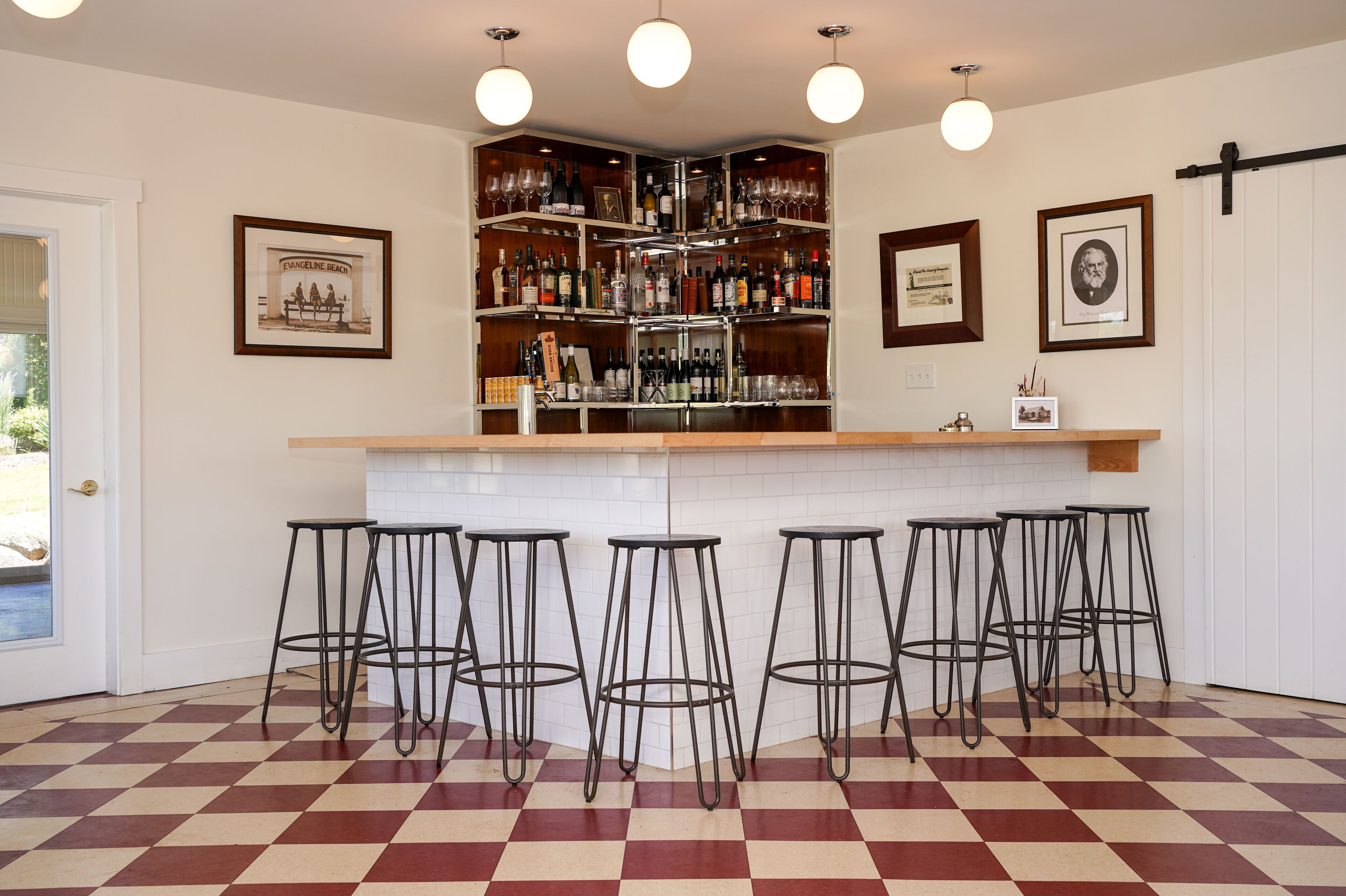 Een bar met een zwart-wit schakeleenvloer, een witte betegelde spatwand en een houten bargedeelte. Er staan vijf zwarte metalen barkrukken en een plank met verschillende flessen en glazen.