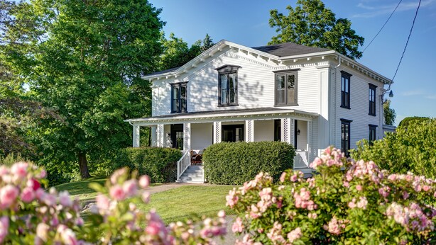 A two-story white house with a front porch, surrounded by lush greenery and flowering plants. - The Evangeline (Grand Pre)
