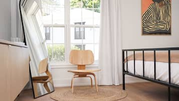 A modern bedroom with a wooden chair, a bed with a black frame, a large window with curtains, and a painting on the wall.