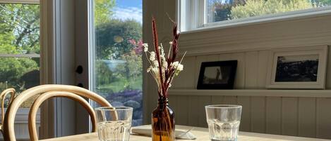 A dining table with two glasses and a vase of dried flowers, set against a window with a view of greenery.