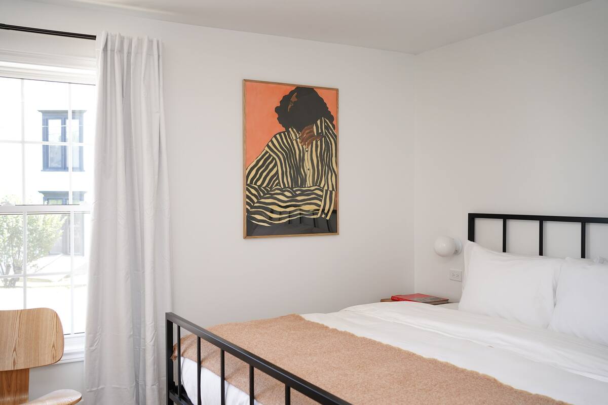 A bedroom with a bed, a window with curtains, a wall-mounted artwork, and a bedside table.