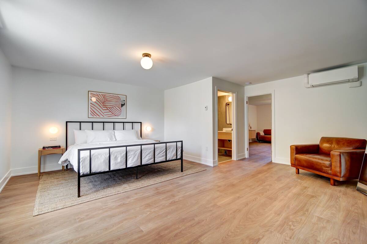 A modern bedroom with a bed, a nightstand, a brown leather sofa, and a hallway leading to another room.