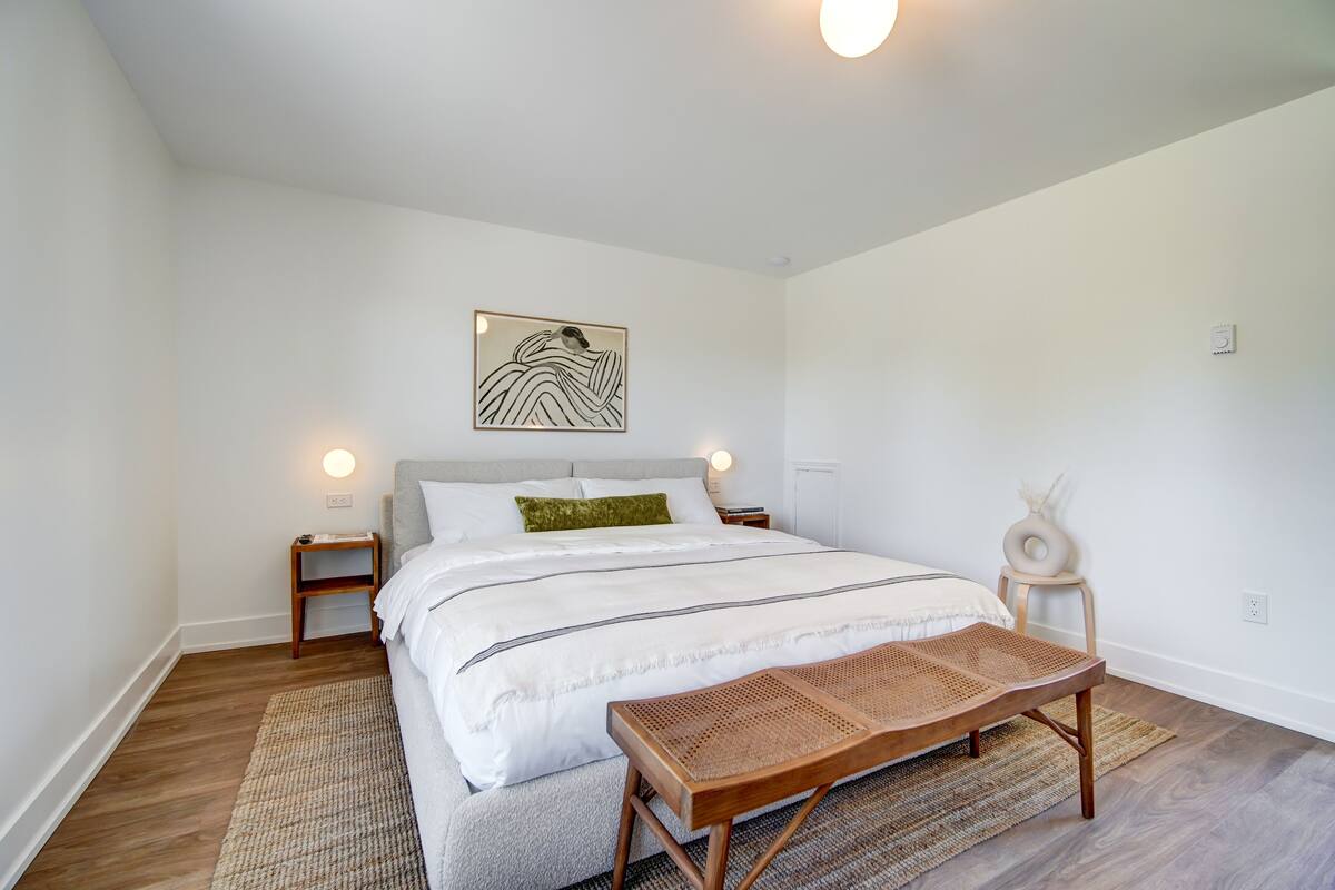 A bedroom with a large bed, a bedside table, a wall-mounted artwork, a small stool, and a woven bench.