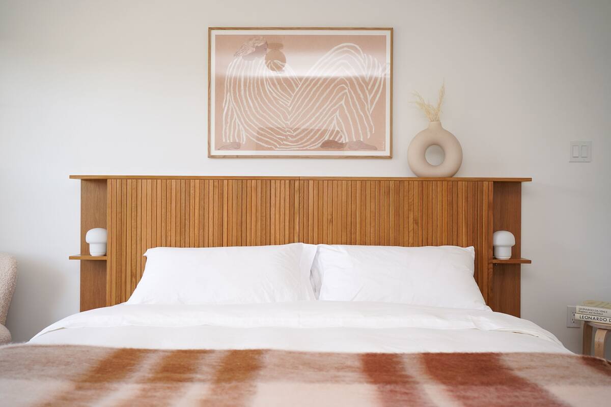 A neatly made bed with white linens, a wooden headboard, and a framed artwork above it.