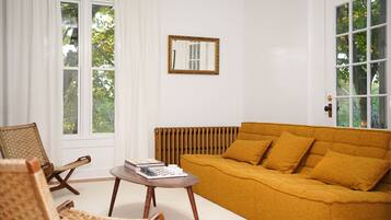 Una sala de estar con un sofá color mostaza, un sillón de mimbre, una mesita y un radiador.