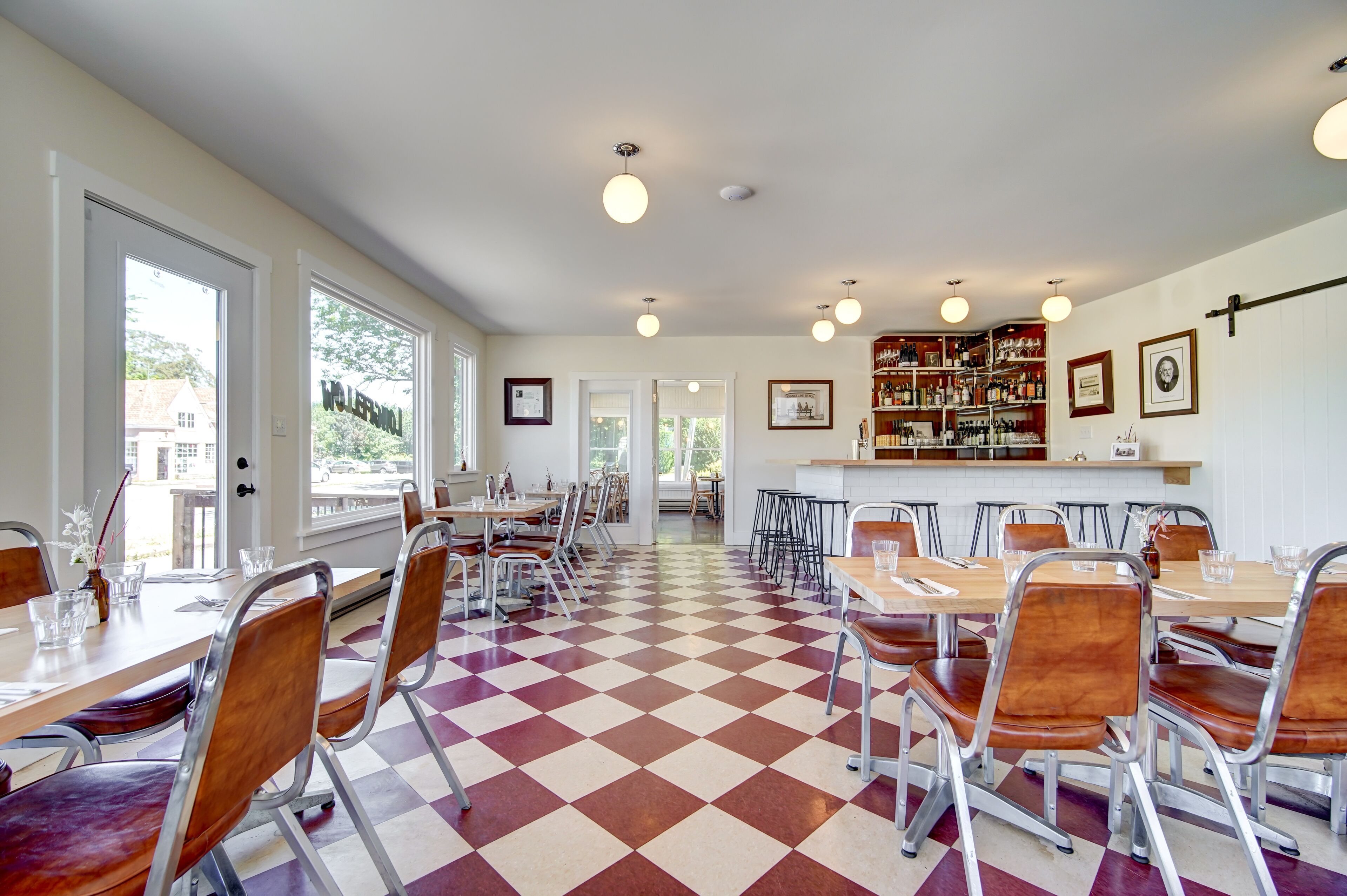 Un espace repas avec un sol à carreaux, des chaises et des tables en métal. On y trouve également un bar avec des étagères remplies de bouteilles et des tableaux encadrés accrochés au mur.