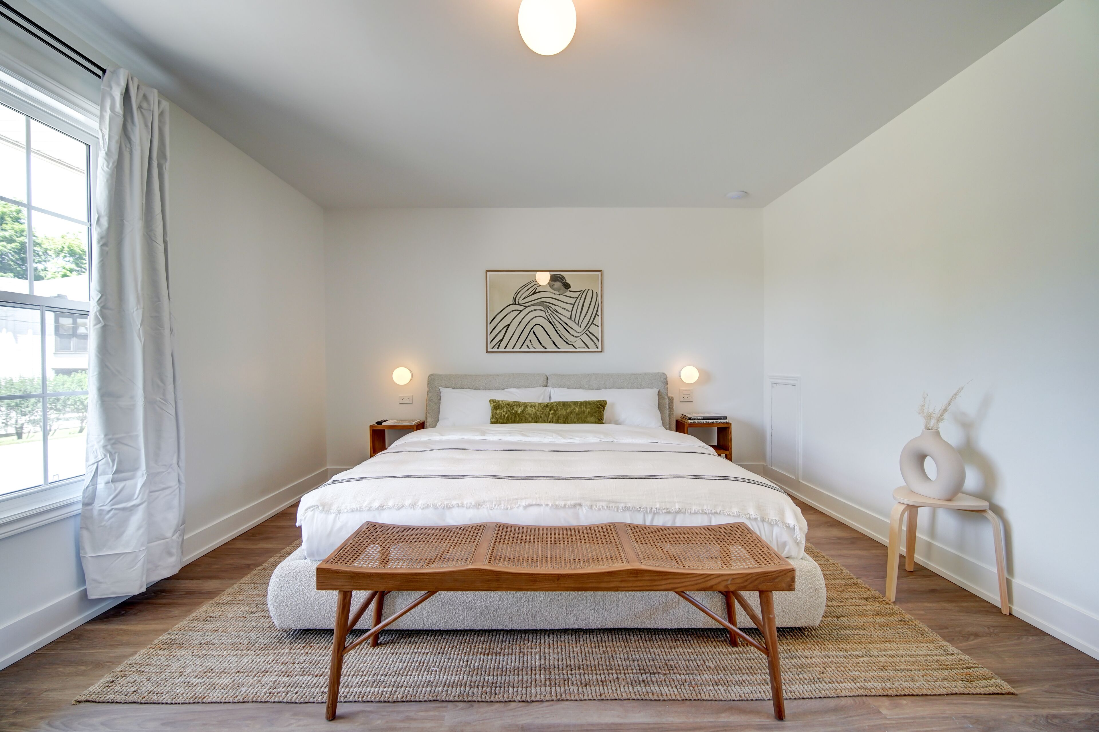 Un dormitorio con una cama grande, una mesita de noche, una obra de arte en la pared y una ventana con cortinas.
