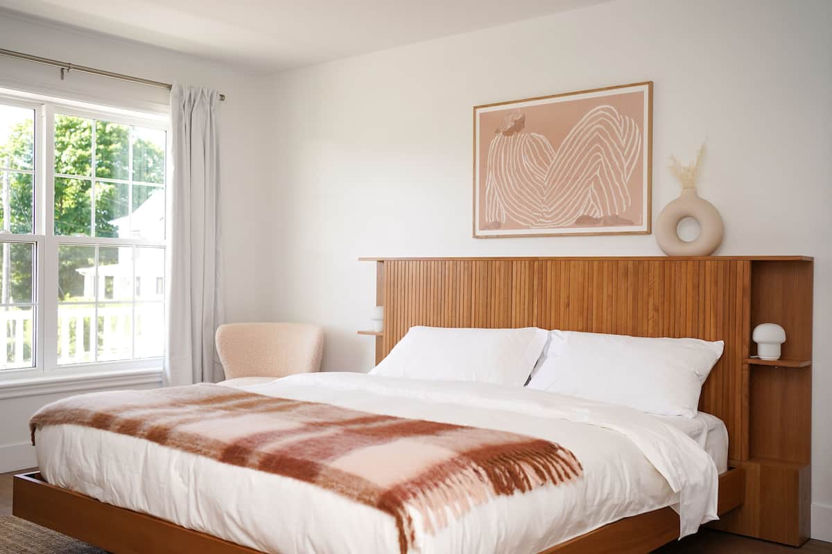 A bedroom with a wooden headboard, a large window with curtains, a bed with white bedding, and a wall-mounted artwork.