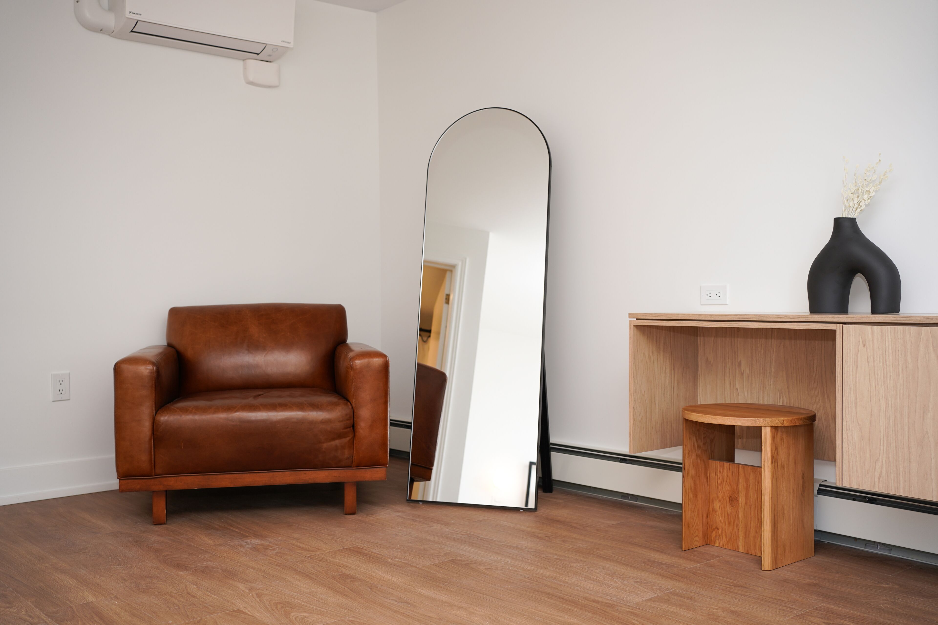 A room with a brown leather sofa, a large mirror, a wooden stool, and a cabinet with a vase.