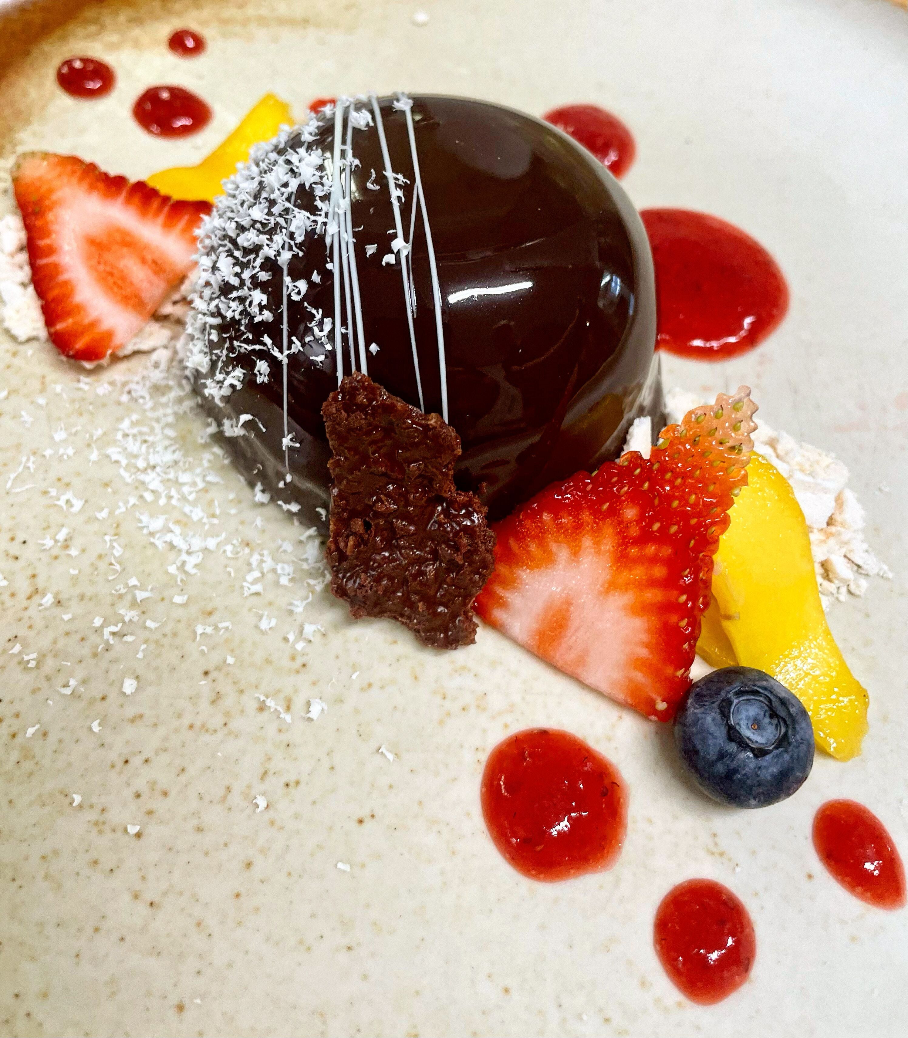 Ein Dessert mit Schokolade, Erdbeeren, Blaubeeren und einem gelben Obst auf einem Teller.