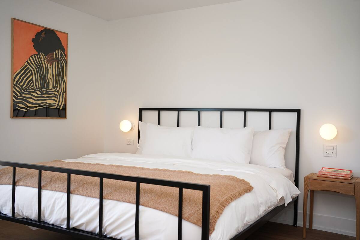 A neatly made bed with white bedding and a brown throw, a bedside table with a book, and a wall-mounted light fixture.