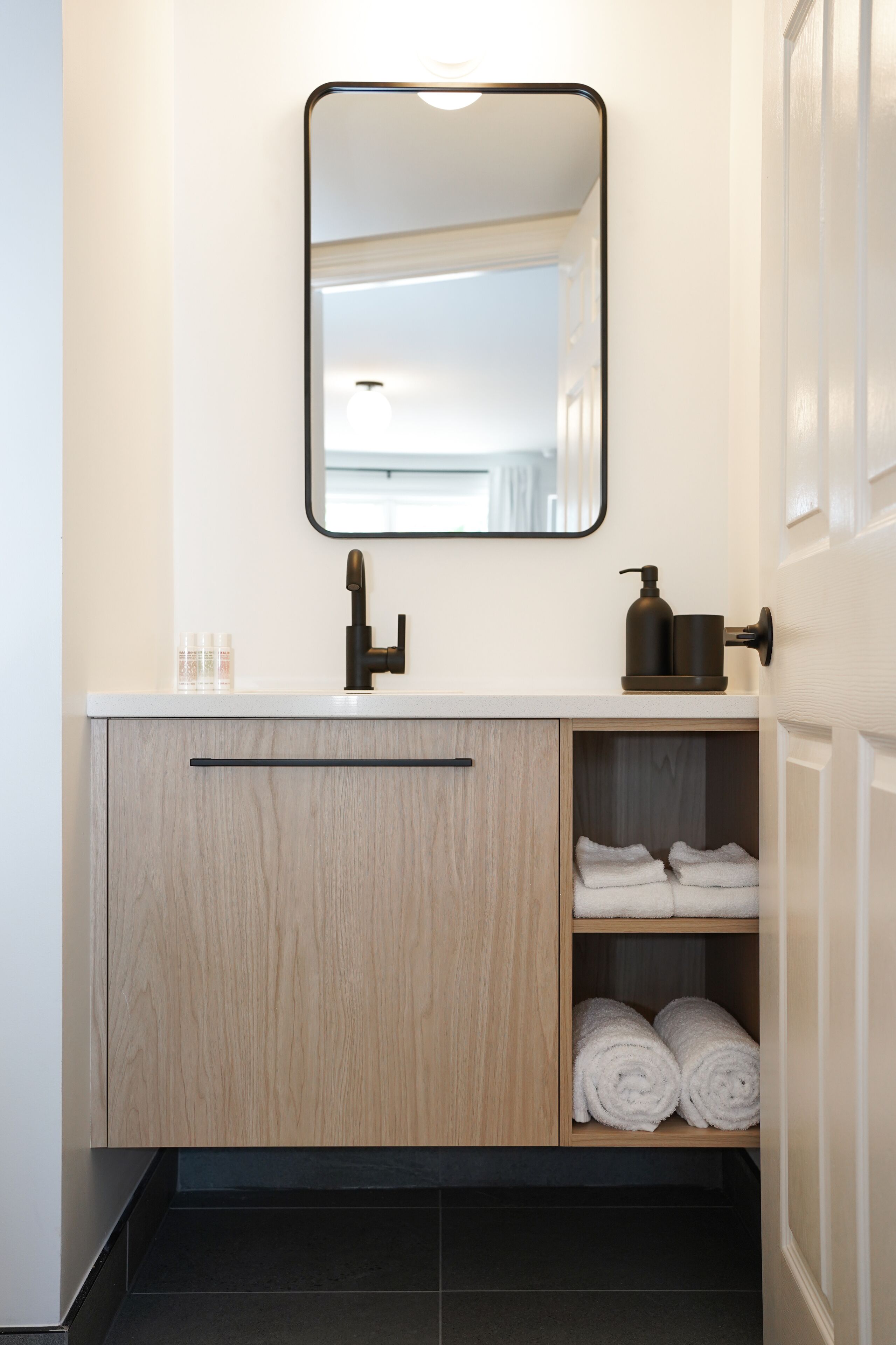 A modern bathroom with a white countertop, a black faucet, a mirror, and a wooden cabinet with towels.